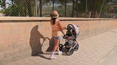 Güneşli bir günde tuğla duvar boyunca bebek arabasıyla gezen sıradan yaz kıyafetleri içinde bir anne. Arka planda ağaçlar ve çitlerle çevrili bir alan yer alıyor..
