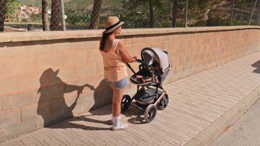Kadın, bebek arabasını tuğla duvarın yanındaki kaldırımdan aşağı itiyor ve sessiz bir yürüyüşün tadını çıkarıyor. Hasır şapka ve günlük yaz kıyafetleri giyiyor..