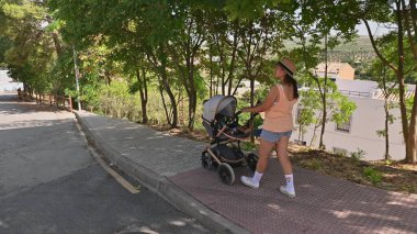 Bir kadın güneşli bir günde yürümekten zevk alır, bebek arabasını ağaçlı kaldırımdan iterek. Sahne, dışarıda huzurlu ve rahatlatıcı bir anı yakalıyor..
