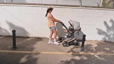 Gündüz kıyafetleri içinde güneşli bir günde bebek arabası süren genç bir anne. Barışçıl bir yürüyüşün tadını çıkarıyor, şapka takıp gülümsüyor..