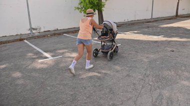 Güneşli bir günde boş bir otoparkta bebek arabasıyla koşan bir kadın. Günlük yaz kıyafetleri ve bir şapka giyiyor..