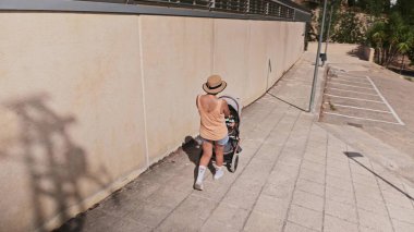 Günlük kıyafetli bir kadın güneşli bir şehir yolunda bebek arabasıyla koşuyor. Olay yeri faaliyet, kentsel yaşam ve sağlıklı, aktif bir yaşam tarzı yayıyor..