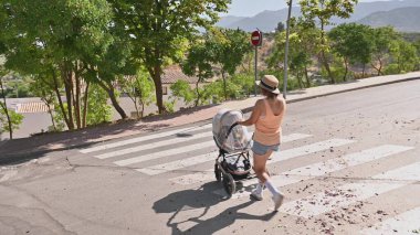 Gündelik yaz kıyafetleri içinde bir kadın güneşli bir günde bir bebek arabasını caddenin karşısına itiyor. Sahne, şehir ortamında sükunet ve açık hava aktivitelerini gösteriyor..