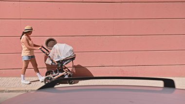 Parlak, güneşli bir günde pembe bir duvarın yanındaki kaldırımda bebek arabasını iten kadın. Şapkalı ve şortlu sıradan yaz kıyafeti..