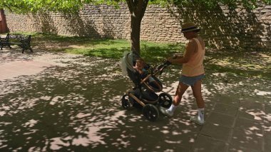 Güneşli bir günde ağaçların gölgesinde yürürken bebek arabasını iten bir anne. Dışarıdaki huzurlu ortamda taş bir duvar ve banklar var..