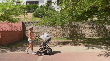 Gündelik yaz kıyafetleri içinde aktif bir kadın güneşli bir günde bebek arabası sürerken koşuyor, taş duvarlı ve yemyeşil yemyeşil huzurlu bir çevrede sağlıklı bir yaşam tarzı sergiliyor..