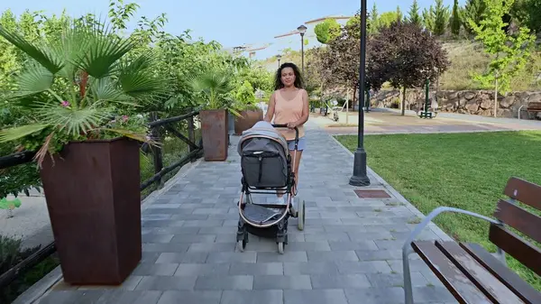 Güneşli bir günde bebeğiyle parkta huzurlu bir yürüyüşün tadını çıkaran bir anne. Etrafı yeşilliklerle çevrili..