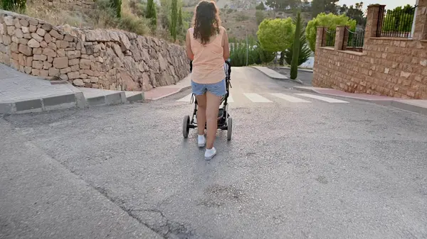 Taş duvarlar ve yeşilliklerle çevrili bir yerleşim yerindeki dik bir tepeden bebek arabasını iten bir kadının arka görüntüsü..