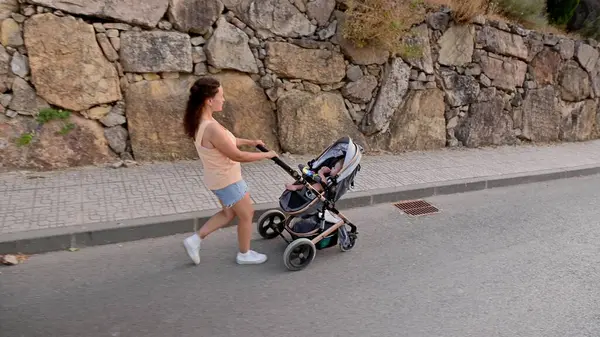 Bir annenin bebeğiyle birlikte arka planda taş bir duvarla sessiz bir cadde boyunca gezindiği görüntüsü..