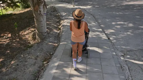 Gündelik yaz kıyafetleri içinde bir kadın gölgeli kaldırımda yürür ve bebek arabasını iter. Şapka ve şort giyiyor, doğada huzurlu bir gezintinin tadını çıkarıyor..