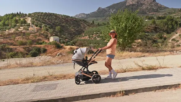 Güneşli bir günde bebek arabasıyla yürüyen mutlu bir anne. Arka planda manzaralı dağlık arazi. Sakin ve açık havada aile bağı..