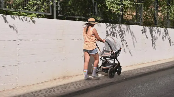 Yaz kıyafetleri giymiş bir kadın güneşli bir günde bebek arabasını iterek şehir kaldırımında yürüyor..