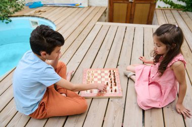 Two children are enjoying a game of chess on a wooden deck by a swimming pool. The boy is making a move while the girl attentively looks on. clipart