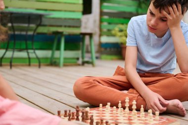 Tahta bir güvertede oturan bir çocuk satranç oyunundaki bir sonraki hamlesini derin derin düşünüyor. Sahne açık havada renkli dekorlarla, odak ve stratejiyi yansıtıyor..