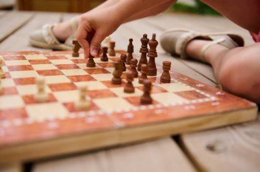 Küçük bir çocuk ahşap bir güvertede satranç oynuyor. Çocuk bir satranç taşını hareket ettiriyor, strateji ve konsantrasyonu gösteriyor..