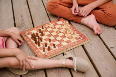 Tahta bir güvertede oturan iki çocuk stratejik bir satranç oynuyorlar. Satranç, konsantrasyonu ve dostça rekabeti teşvik eder..