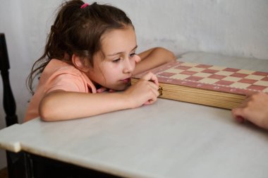 Genç bir kız içeride bir masa oyunu oynarken derin düşüncelere dalmış ve odaklanmış görünüyor. Bir sonraki hamlesini düşünerek başını masaya yaslar..