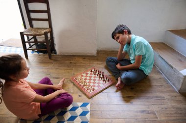 İki çocuk evde satranç oynuyor, yerde oturuyor, strateji ve öğrenmeye odaklanıyor. Eleştirel düşünmeyi teşvik eden eğlenceli ve eğitici bir iç etkinlik..