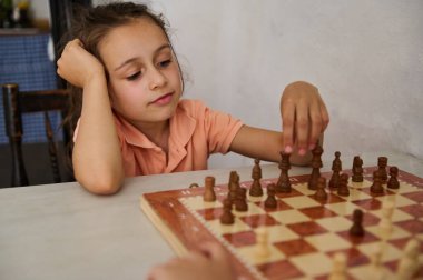Turuncu tişörtlü genç bir kız satranç oynuyor ve satranç tahtasına odaklanmış bir ifadeyle bir parça yerleştiriyor..