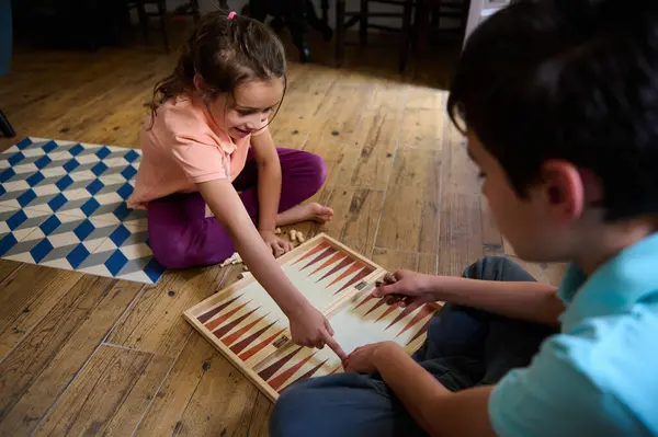 stock image Two children engaged in playing a board game on a wooden floor, creating joyful moments of fun and learning while bonding together.