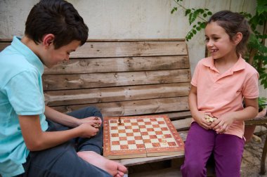 İki çocuk dışarıda satranç oynuyor, tahta bir bankta oturuyor, gülümsüyor ve oyundan zevk alıyorlar. Görüntü, eğlence, strateji ve eğlenceyi yakalar..