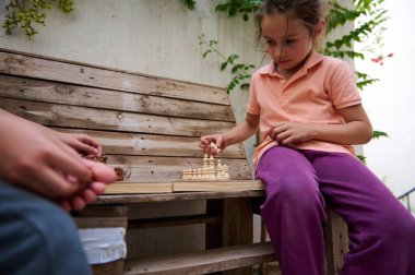 Kız dışarıda tahta bir bankta satranç oynuyor, eğlenceli ve eğitici bir oyun oynuyor. Stratejik düşünme ve öğrenme ile meşgul doğal bir açık hava ortamında bir arkadaş ile.