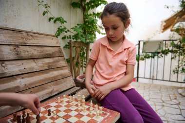 Genç bir kız, bir sonraki hamlesine odaklanır dışarıda, ahşap bir bankta satranç oynarken, bahçe ortamında sessiz ve stratejik bir oyunun tadını çıkarırken..