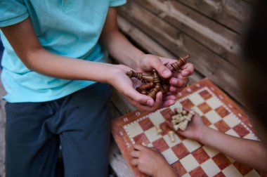 Çocuklar dışarıda satranç oynuyor, stratejiye odaklanıyor ve arkadaşça bir oyun oynuyorlar. Satranç taşlarını tutan eller, konsantrasyon ve zihinsel becerileri vurguluyor.