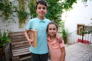 İki kardeş açık havada satranç tahtası tutarak mutlu bir şekilde poz veriyorlar. Huzurlu bir bahçe ortamında aile bağlarını ve neşeli anları temsil ederler..