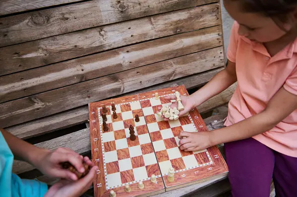 İki çocuk ahşap bir bankta satranç oynuyor. Stratejik bir oyuna dalmış, dışarıda eğleniyor ve öğreniyor..