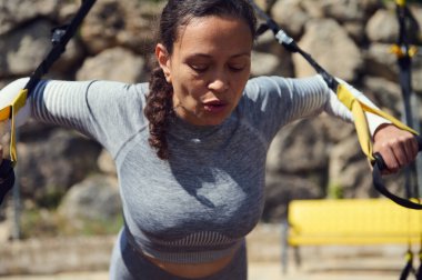 Kaslı bir kadın açık havada süspansiyon egzersizleri yapıyor. Güç eğitimini ve doğal ortamdaki uygunluğa olan bağlılığını vurguluyor..