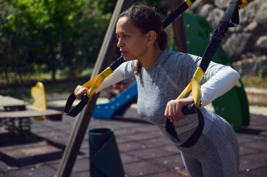 Kararlı genç bir kadın, açık havada süspansiyon kayışı eğitimi alıyor. Doğal park ortamında spor yapmaya odaklanıyor ve kendini spora veriyor..