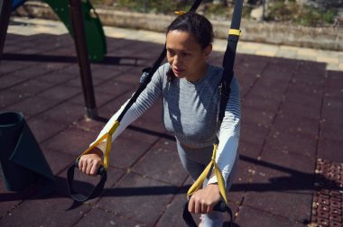 Kararlı bir kadın açık havada TRX süspansiyon kayışlarıyla çalışıyor güneşli bir park ortamında güç ve zindelik gösterisi yapıyor. Egzersiz ve motivasyon kavramları için mükemmel..
