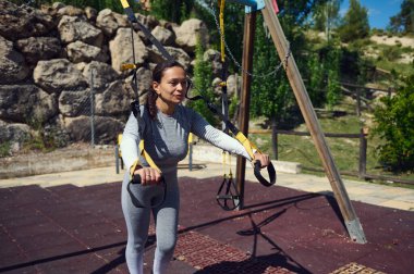 Açık havada direnç bantları kullanan kararlı bir kadın güneşli bir günde doğal park ortamında spor ve dayanıklılık eğitimini destekliyor..