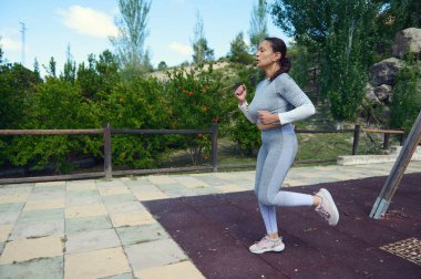 Parkta koşan atletik giysili bir kadın aktif bir yaşam tarzını benimsiyor. Doğal ortamdaki spor, sağlık ve uygunluğu vurgular.