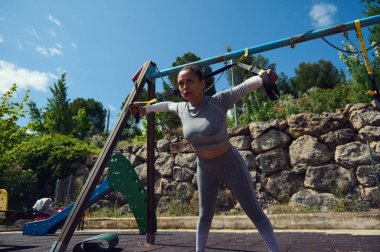 Bir kadın, açık hava oyun alanında askılarla egzersiz yapıyor. Doğal ortamda spor, güç ve sağlığa vurgu yapmak.