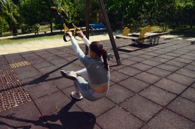 Güneşli bir parkta açık hava süspansiyon eğitimi alan bir kadın denge ve güç gösterisi sergiliyor. Kendini spora adaması bu dinamik egzersiz sahnesinde açıkça görülüyor..