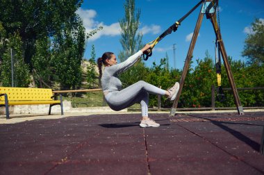 Parkta TRX kayışlarıyla çalışan formda bir kadın açık havada denge, güç ve zindelik gösteriyor..