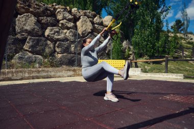 Aktif kadın dışarıda TRX egzersizi yapıyor. Güç ve denge gösterisi yapıyor. Kayalar ve ağaçlarla doğal bir ortamda kurulmuş, fitness ve açık hava yaşam tarzını ön plana çıkarıyor..