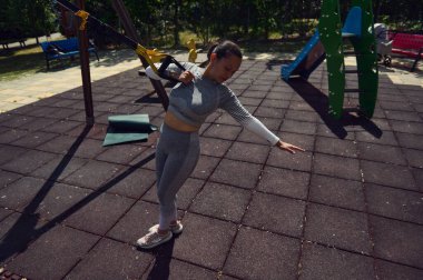TRX kayışlarıyla parkta egzersiz yapan bir kadın. Spor meraklısı güneşli gökyüzünün altında vücut çalışıp güç toplamaya çalışıyor..