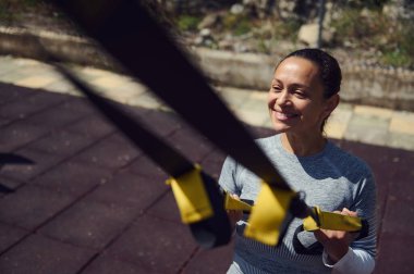 Aktif bir kadın TRX süspansiyon antrenörü kullanıyor, spor yaparken gülümsüyor. Güneşli bir park ortamında spor ve sağlıklı yaşam tarzını kucaklamak.