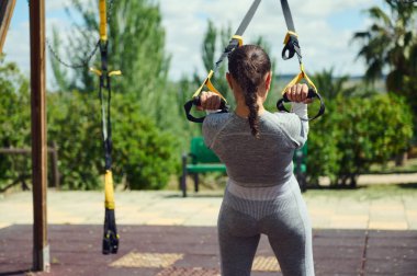 TRX kayışlarıyla dışarıda egzersiz yapan bir kadının arka görüntüsü. Odak ve kararlılığı yemyeşil bir park ortamında yakalar.