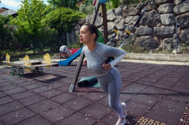 Açık havada TRX kayışlarıyla egzersiz yapan odaklanmış bir kadın. Vücut yapısını somutlaştıran, sağlıklı ve kararlı bir yaşam sürüyor..
