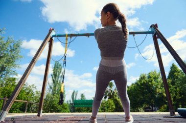 Spor kıyafetleri içinde bir kadın çocuk parkında, açık gökyüzünün altında antrenmana hazırlanıyor..