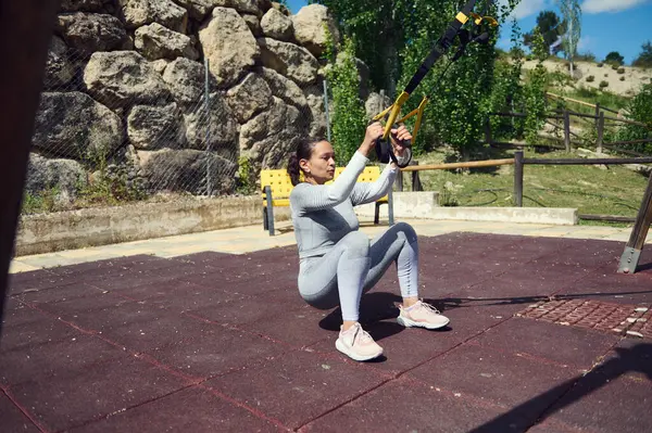 stock image Young woman engaging in TRX workout in an outdoor park setting, emphasizing strength and fitness training on a sunny day.