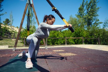 Güneşli bir spor salonunda TRX kayışlarıyla egzersiz yapan bir kadın güç, denge ve atletik kararlılık sergiliyor..