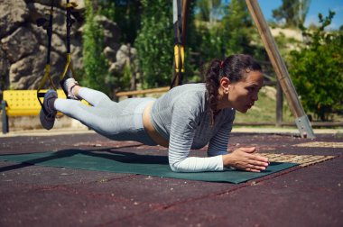 Açık hava parkı ortamında TRX kayışlarıyla tahta egzersizi yapan bir kadın. Spor ve dayanıklılık eğitimine odaklanmış durumda. Doğal ortamdaki egzersizlerin tadını çıkarıyor..