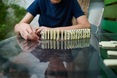 Bir çocuk dikkatli bir konsantrasyon ve stratejik düşünceyi vurgulamak için ustalıkla cam bir masaya domino taşları yerleştirir. Odak noktası dominoların yansıması ve konumu..