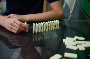 Bir kişi, strateji ve hassasiyete odaklanarak, bir cam masaya domino taşlarını yerleştirir. Sahne, oyundaki beklenti ve beceriyi yakalıyor..