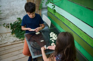Dışarıda bahçede domino oynayan iki çocuk, yeşil paneller ve bitkilerle çevrili. Oyuna odaklanırlar, rahat bir öğleden sonranın tadını çıkarırlar..